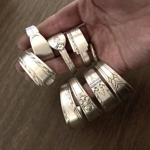 May include: A collection of silver spoon bracelets with various ornate designs. The bracelets are stacked on top of each other and are held in a hand.