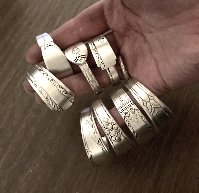 May include: A collection of silver spoon bracelets with various ornate designs. The bracelets are stacked on top of each other and are held in a hand.