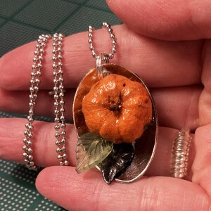May include: A silver chain necklace with an orange pumpkin charm and green and black leaves. The charm is attached to a silver oval pendant.