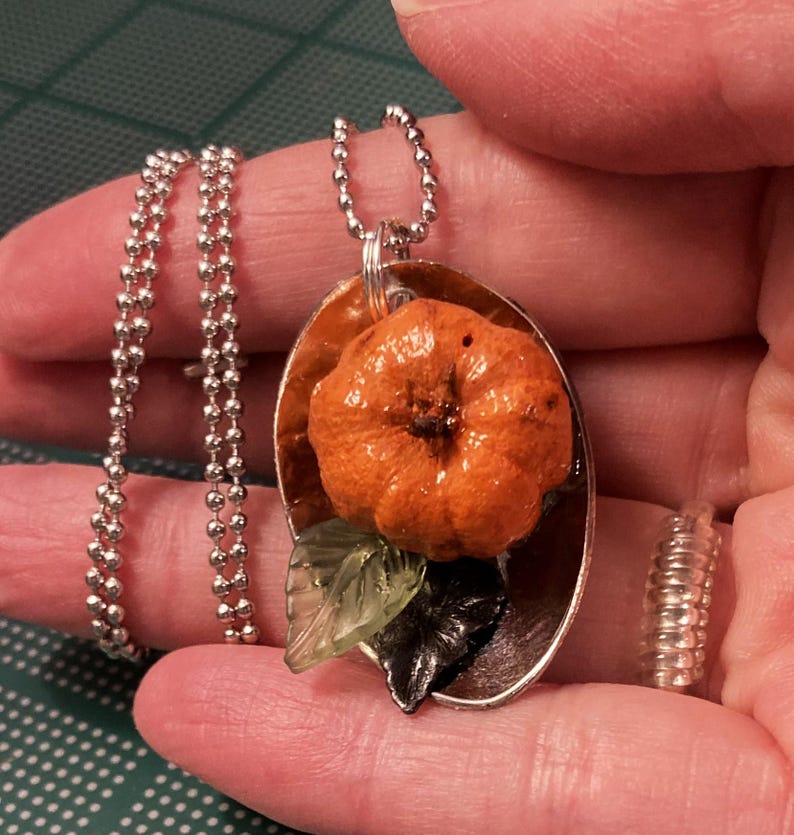 May include: A silver chain necklace with an orange pumpkin charm and green and black leaves. The charm is attached to a silver oval pendant.