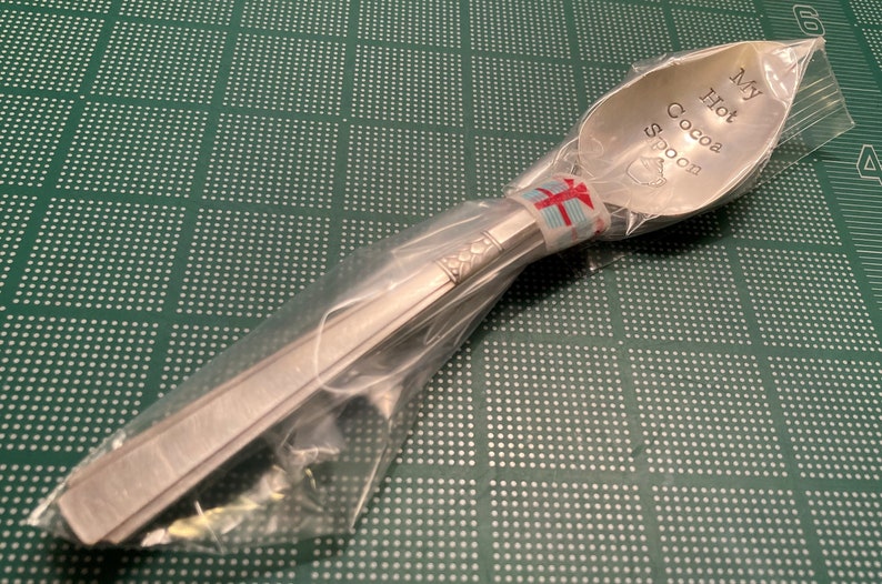May include: A silver spoon with the words "My Hot Cocoa Spoon" engraved on the bowl. The spoon is wrapped in clear plastic and tied with a red and white ribbon.