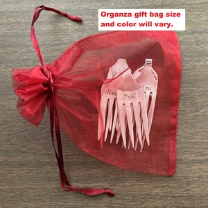 May include: A red organza gift bag with a drawstring closure. The bag contains a set of six silver hair forks with engraved names, including "Hav", "Brie", and "Jeu".