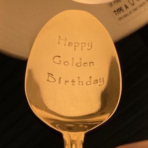 May include: A gold-colored spoon with the words "Happy Golden Birthday" engraved on the bowl. The spoon has a classic teardrop shape and a decorative handle. The text is centered and in a simple, readable font.
