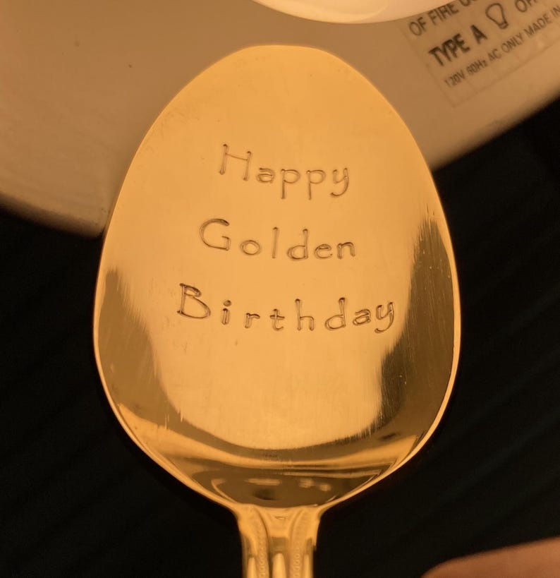 May include: A gold-colored spoon with the words "Happy Golden Birthday" engraved on the bowl. The spoon has a classic teardrop shape and a decorative handle. The text is centered and in a simple, readable font.