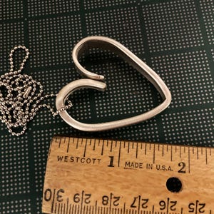 May include: A silver heart-shaped pendant necklace with a ball chain. The pendant is about 1 1/2 inches wide and is made from two pieces of metal that are bent into a heart shape. The pendant is hanging from a silver ball chain.