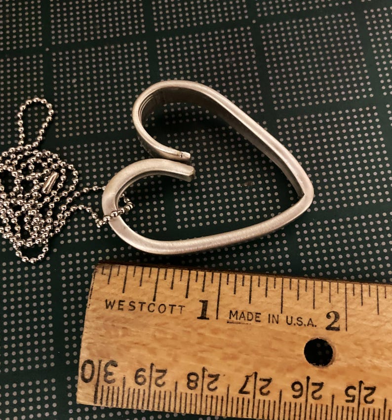 May include: A silver heart-shaped pendant necklace with a ball chain. The pendant is about 1 1/2 inches wide and is made from two pieces of metal that are bent into a heart shape. The pendant is hanging from a silver ball chain.