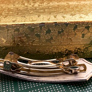 May include: A gold-toned metal hair clip with a decorative design. The clip is sitting on a green surface with a white polka dot pattern.