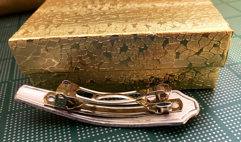 May include: A gold-toned metal hair clip with a decorative design. The clip is sitting on a green surface with a white polka dot pattern.