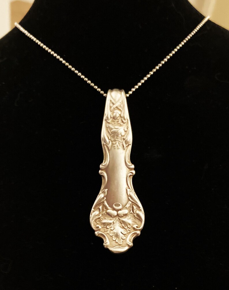 Puede incluir: Un collar de plata con un colgante hecho de una cuchara de plata vintage. La cuchara tiene un dise&ntilde;o adornado con un motivo de hojas.
