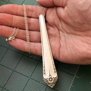 May include: A silver pendant necklace made from a repurposed spoon. The pendant is a long, slender shape with a floral design at the bottom. The chain is delicate and silver, with a clasp. The necklace is held in a hand.