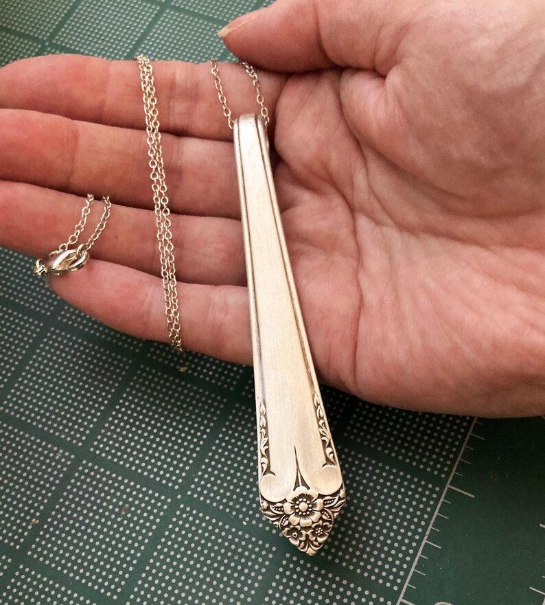 May include: A silver pendant necklace made from a repurposed spoon. The pendant is a long, slender shape with a floral design at the bottom. The chain is delicate and silver, with a clasp. The necklace is held in a hand.