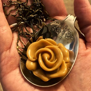 May include: A gold-toned rose pendant necklace with a brown rose charm. The rose charm is sitting on a silver spoon.