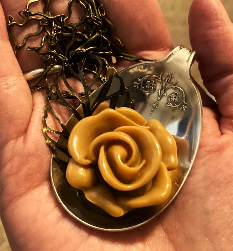 May include: A gold-toned rose pendant necklace with a brown rose charm. The rose charm is sitting on a silver spoon.