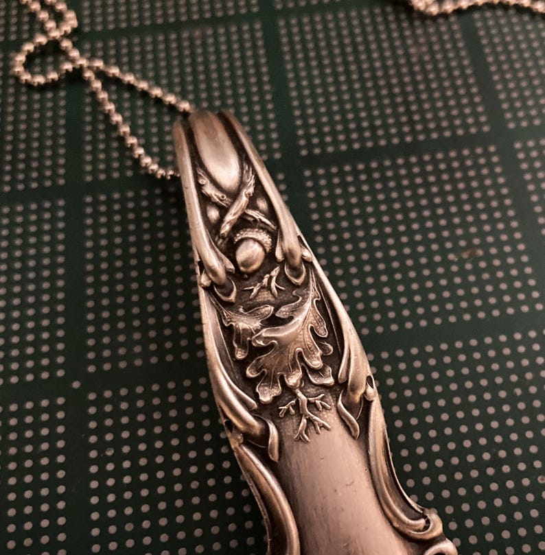 May include: Silver spoon pendant necklace with an ornate design featuring an acorn and oak leaves. The spoon is attached to a silver chain.