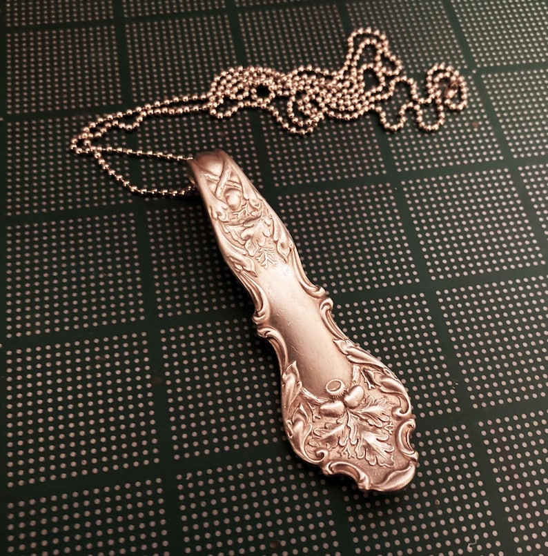 Puede incluir: Un collar de cuchara de plata con un dise&ntilde;o adornado con hojas y flores. La cuchara est&aacute; unida a una cadena de plata.