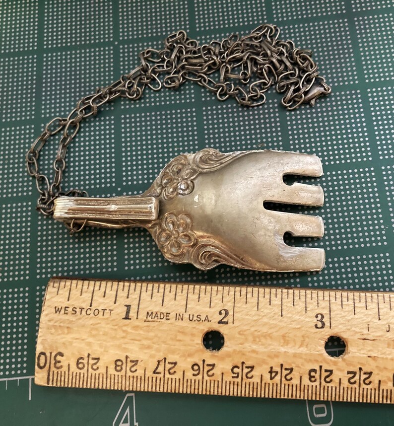 May include: Silver fork necklace with a chain. The fork is about 2 inches long and has a decorative design. The chain is silver and about 18 inches long.