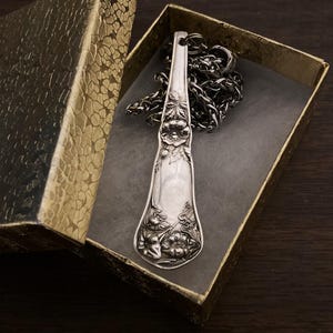 May include: A silver spoon necklace with a floral design. The spoon is hanging in a small, gold gift box.