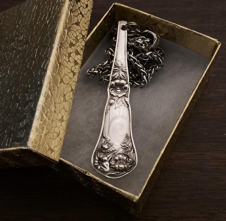 May include: A silver spoon necklace with a floral design. The spoon is hanging in a small, gold gift box.