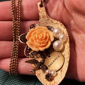 May include: A spoon necklace with a peach-colored rose, white pearls, a bird charm, and a gold tag that says "BALANCE".