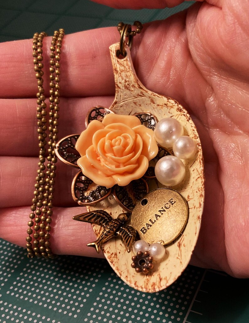 May include: A spoon necklace with a peach-colored rose, white pearls, a bird charm, and a gold tag that says "BALANCE".