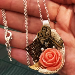 May include: A silver chain necklace with a pendant made from a repurposed spoon. The spoon is decorated with a key, a heart, lace, a rose, and a gold leaf.