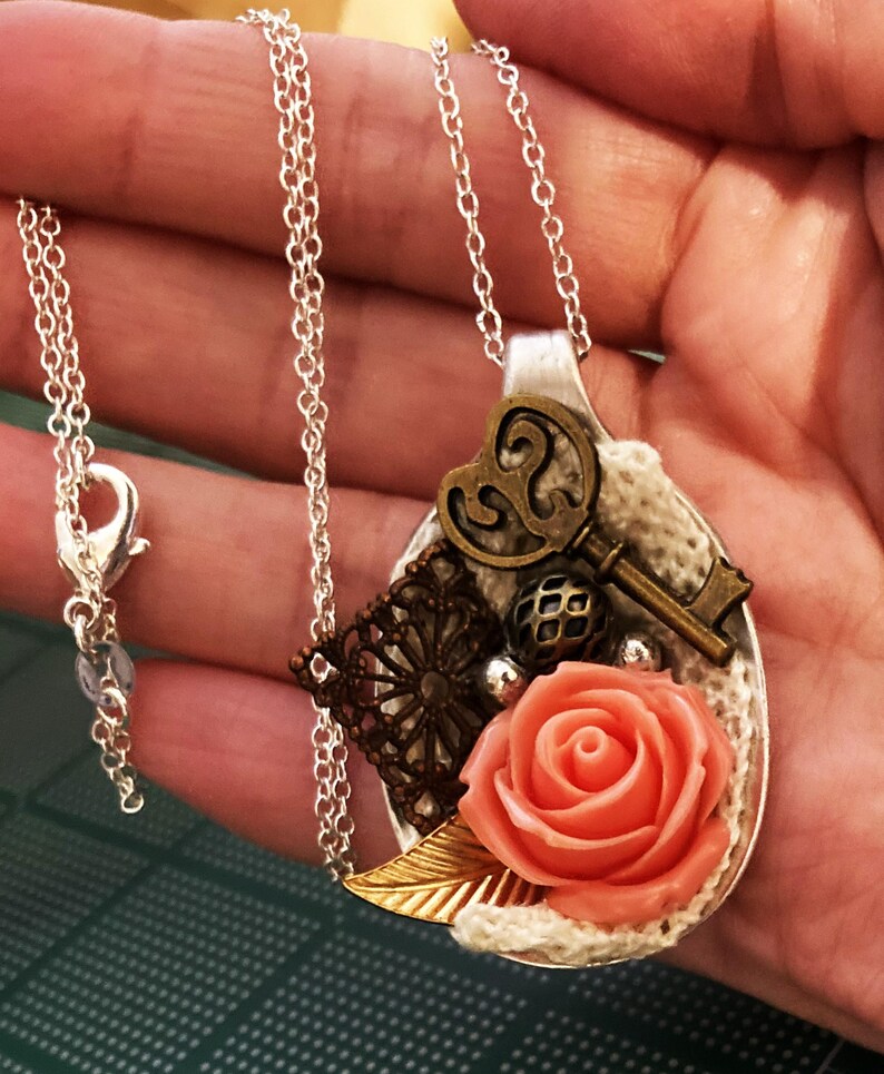 May include: A silver chain necklace with a pendant made from a repurposed spoon. The spoon is decorated with a key, a heart, lace, a rose, and a gold leaf.