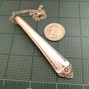 May include: A silver pendant necklace made from a repurposed spoon. The spoon's handle is the focal point, with a floral design at the end. A silver chain and clasp are attached. A quarter coin is next to the pendant.