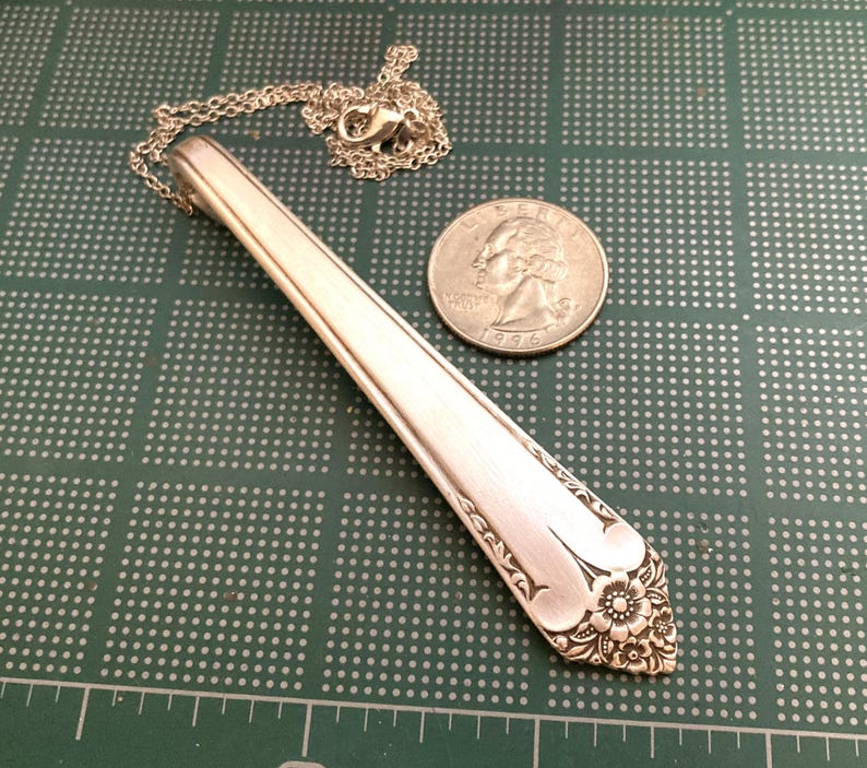 May include: A silver pendant necklace made from a repurposed spoon. The spoon's handle is the focal point, with a floral design at the end. A silver chain and clasp are attached. A quarter coin is next to the pendant.