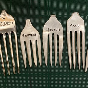 May include: Six silver forks with different cheese names engraved on them: Bleu, Edam, Gruyere, Havarti, Goat, and Brie.