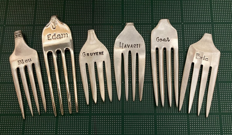 May include: Six silver forks with different cheese names engraved on them: Bleu, Edam, Gruyere, Havarti, Goat, and Brie.