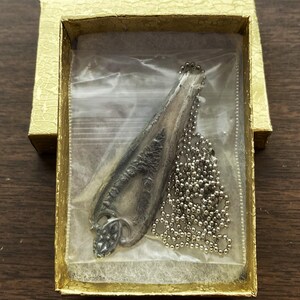 May include: A silver pendant necklace in a gold box. The pendant is shaped like a long, thin leaf with a detailed design. The chain is a silver ball chain.