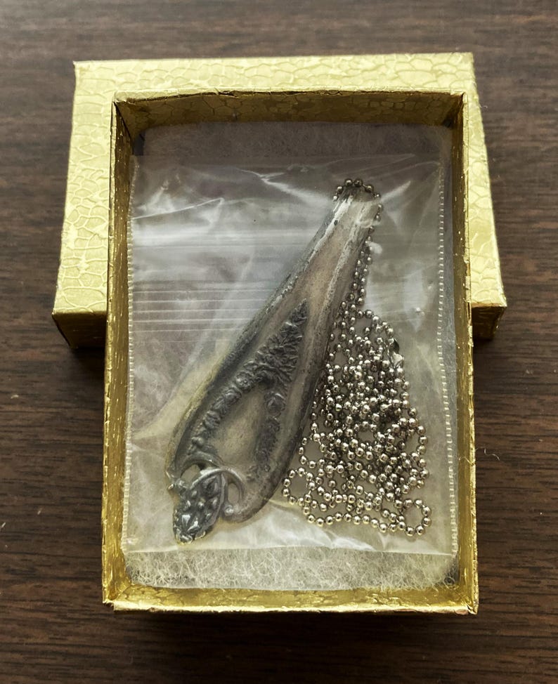 May include: A silver pendant necklace in a gold box. The pendant is shaped like a long, thin leaf with a detailed design. The chain is a silver ball chain.