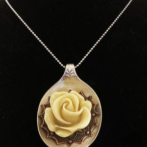 May include: A silver spoon necklace with a white rose charm. The spoon has a decorative edge and the rose is centered on the spoon.