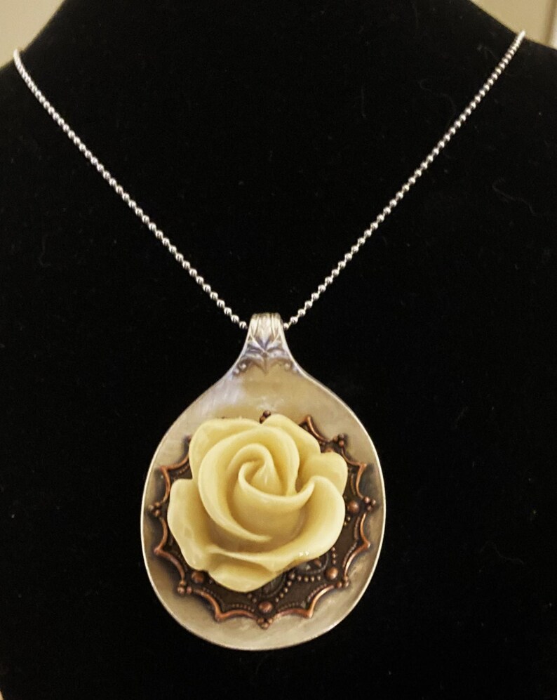 May include: A silver spoon necklace with a white rose charm. The spoon has a decorative edge and the rose is centered on the spoon.