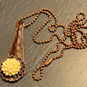 May include: A brown spoon pendant necklace with a white flower charm. The necklace is made with a brown ball chain.