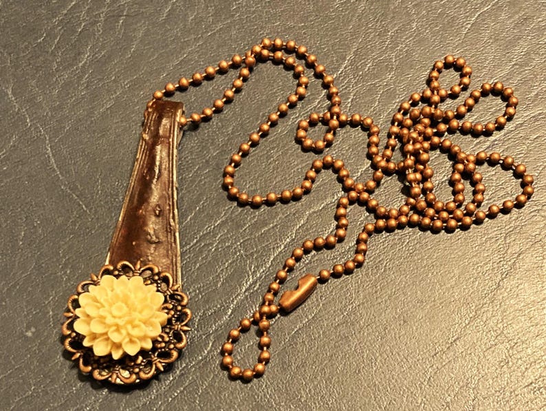May include: A brown spoon pendant necklace with a white flower charm. The necklace is made with a brown ball chain.