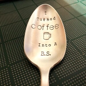 Pu&ograve; includere: Un cucchiaio d'argento con la scritta "I Turned Coffee Into A B.S." incisa sulla conca del cucchiaio. Un piccolo disegno di una tazza di caff&egrave; &egrave; tra le parole "Coffee" e "Into". Il cucchiaio &egrave; su una superficie verde e bianca a pois.