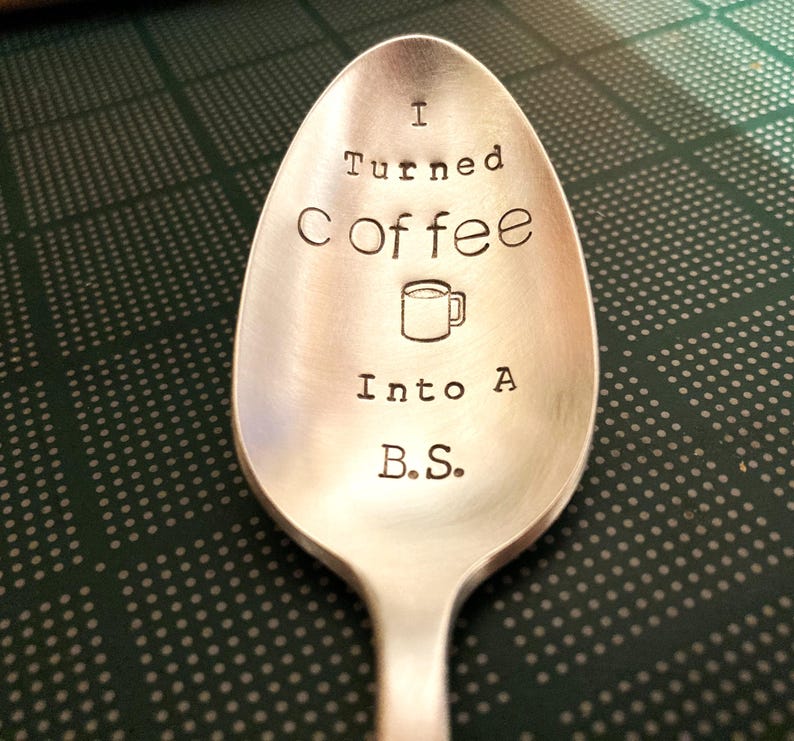 Pu&ograve; includere: Un cucchiaio d'argento con la scritta "I Turned Coffee Into A B.S." incisa sulla conca del cucchiaio. Un piccolo disegno di una tazza di caff&egrave; &egrave; tra le parole "Coffee" e "Into". Il cucchiaio &egrave; su una superficie verde e bianca a pois.
