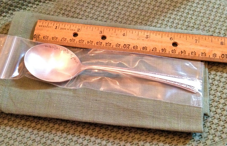 May include: A silver spoon with a personalized inscription that reads "mackenzie's spour". The spoon is packaged in a clear plastic bag.