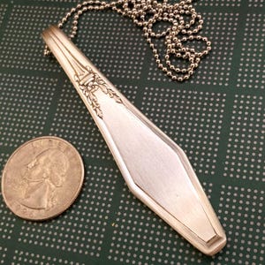 Puede incluir: Un collar de cuchara de plata con un diseño floral decorativo en el mango. La cuchara se ha remodelado en forma de lágrima y está unida a una cadena de bolas de plata.