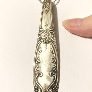 May include: A silver spoon handle necklace with an ornate design. The handle is long and narrow with a floral pattern.