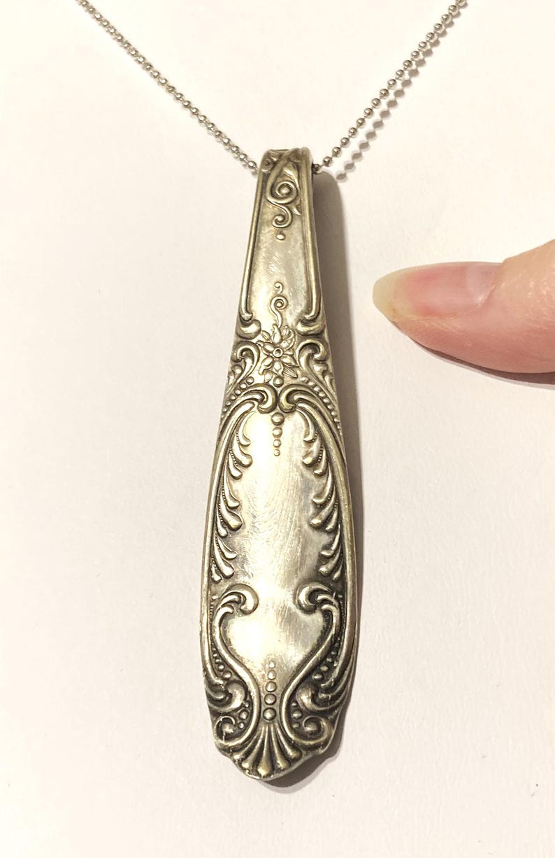 May include: A silver spoon handle necklace with an ornate design. The handle is long and narrow with a floral pattern.