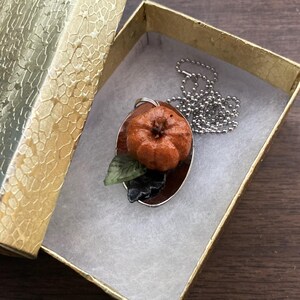 May include: A silver chain necklace with a pendant featuring a small orange pumpkin with a green leaf. The pendant is on a brown background.