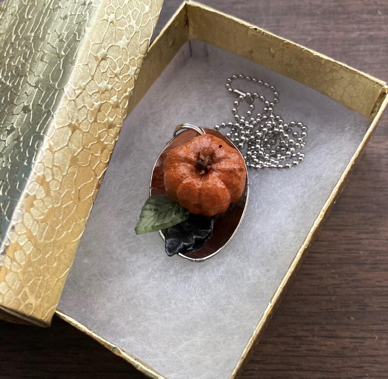 May include: A silver chain necklace with a pendant featuring a small orange pumpkin with a green leaf. The pendant is on a brown background.