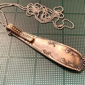 May include: A silver spoon necklace with a decorative design on the handle. The spoon is attached to a silver chain.