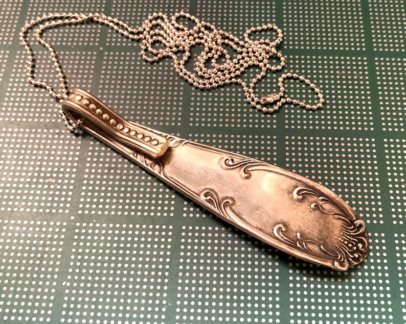 May include: A silver spoon necklace with a decorative design on the handle. The spoon is attached to a silver chain.