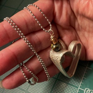 May include: A silver heart-shaped pendant necklace with a silver ball chain. The pendant is made from a repurposed spoon and features a decorative design. The necklace also includes a multi-colored bead charm.