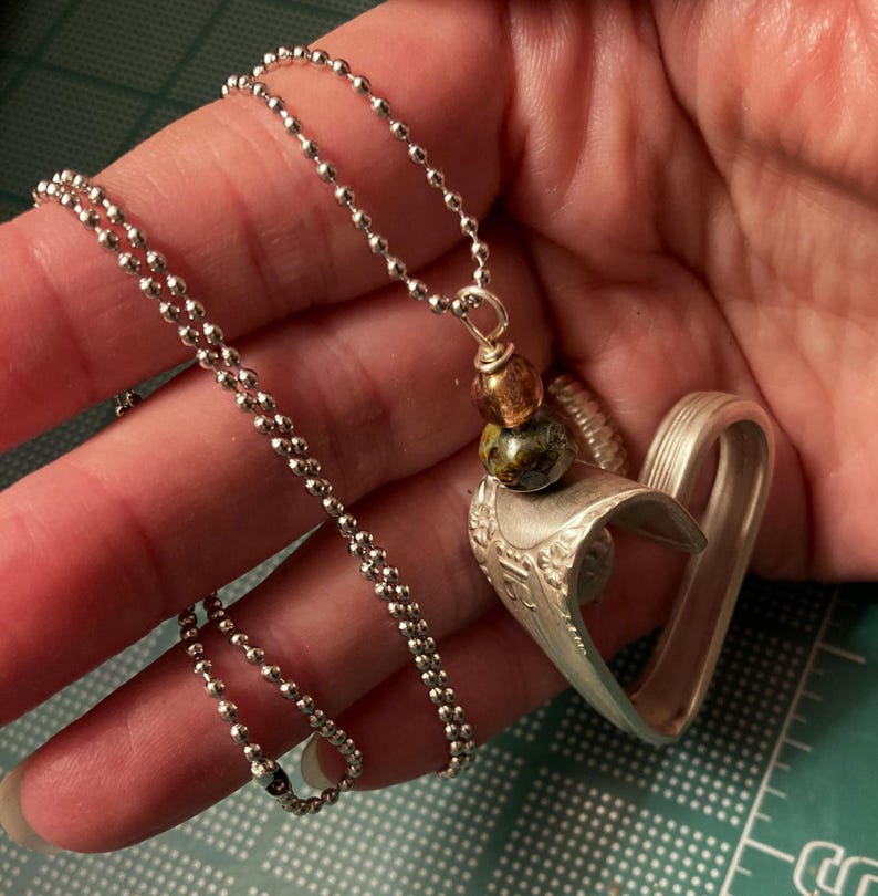 May include: A silver heart-shaped pendant necklace with a silver ball chain. The pendant is made from a repurposed spoon and features a decorative design. The necklace also includes a multi-colored bead charm.