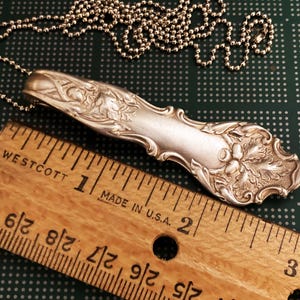 Puede incluir: Un collar de cuchara de plata con un dise&ntilde;o ornamentado que presenta un racimo de bayas y hojas. La cuchara mide aproximadamente 5 cm de largo y est&aacute; unida a una cadena de plata.