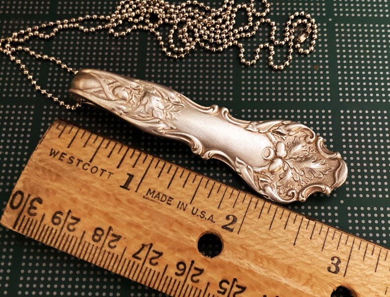 Puede incluir: Un collar de cuchara de plata con un dise&ntilde;o ornamentado que presenta un racimo de bayas y hojas. La cuchara mide aproximadamente 5 cm de largo y est&aacute; unida a una cadena de plata.